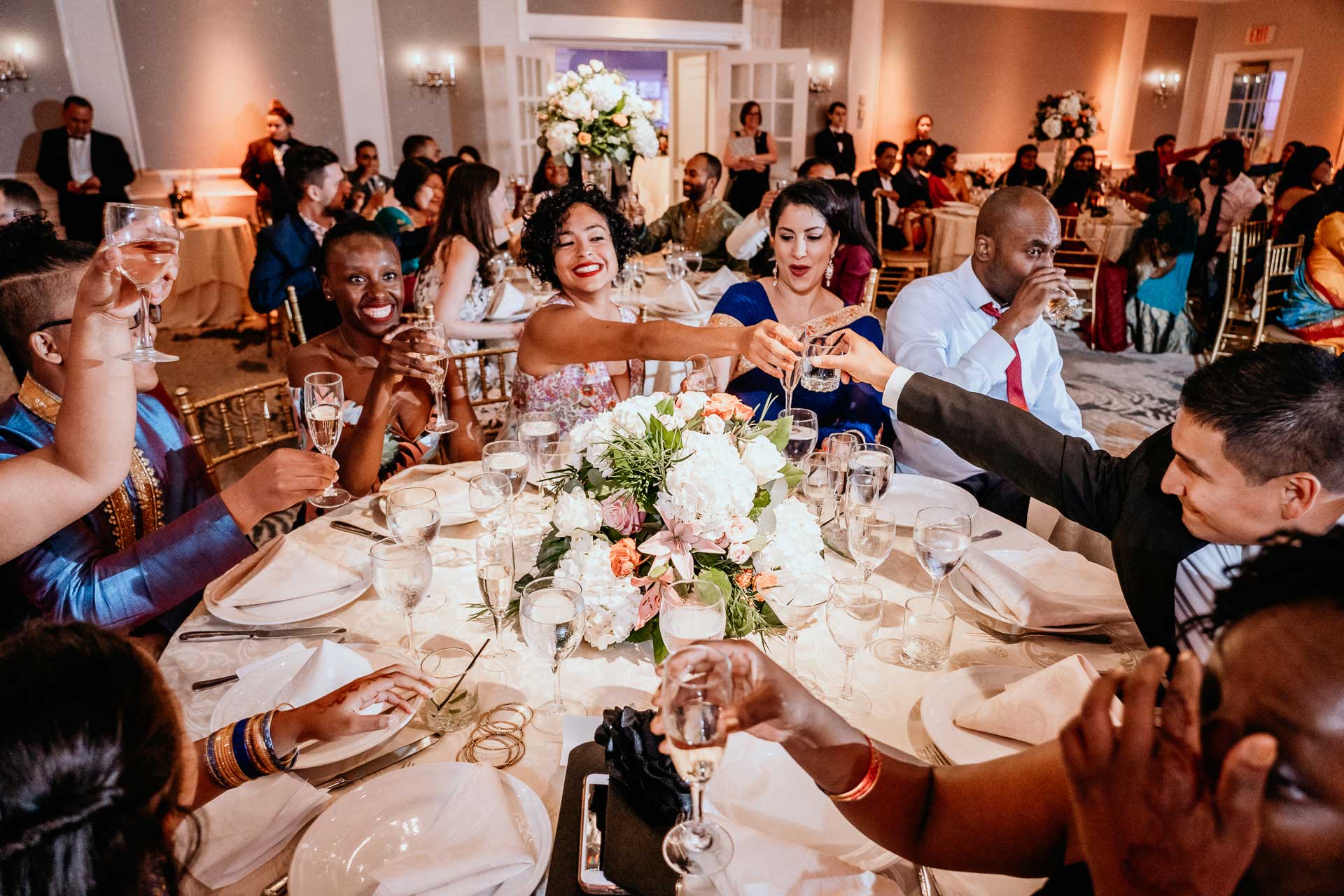 brooklake-Indian-Wedding-photographer-062 A group of people dressed formally, seated around a decorated round table, raise glasses in a toast at a lively NJ Teen Events celebration.