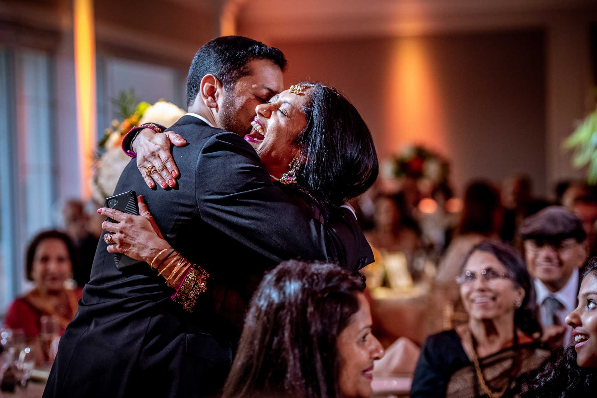 brooklake-Indian-Wedding-photographer-063 A man and woman share a joyful hug at an indoor NJ Teen Events gathering, surrounded by seated guests who are smiling and watching them.