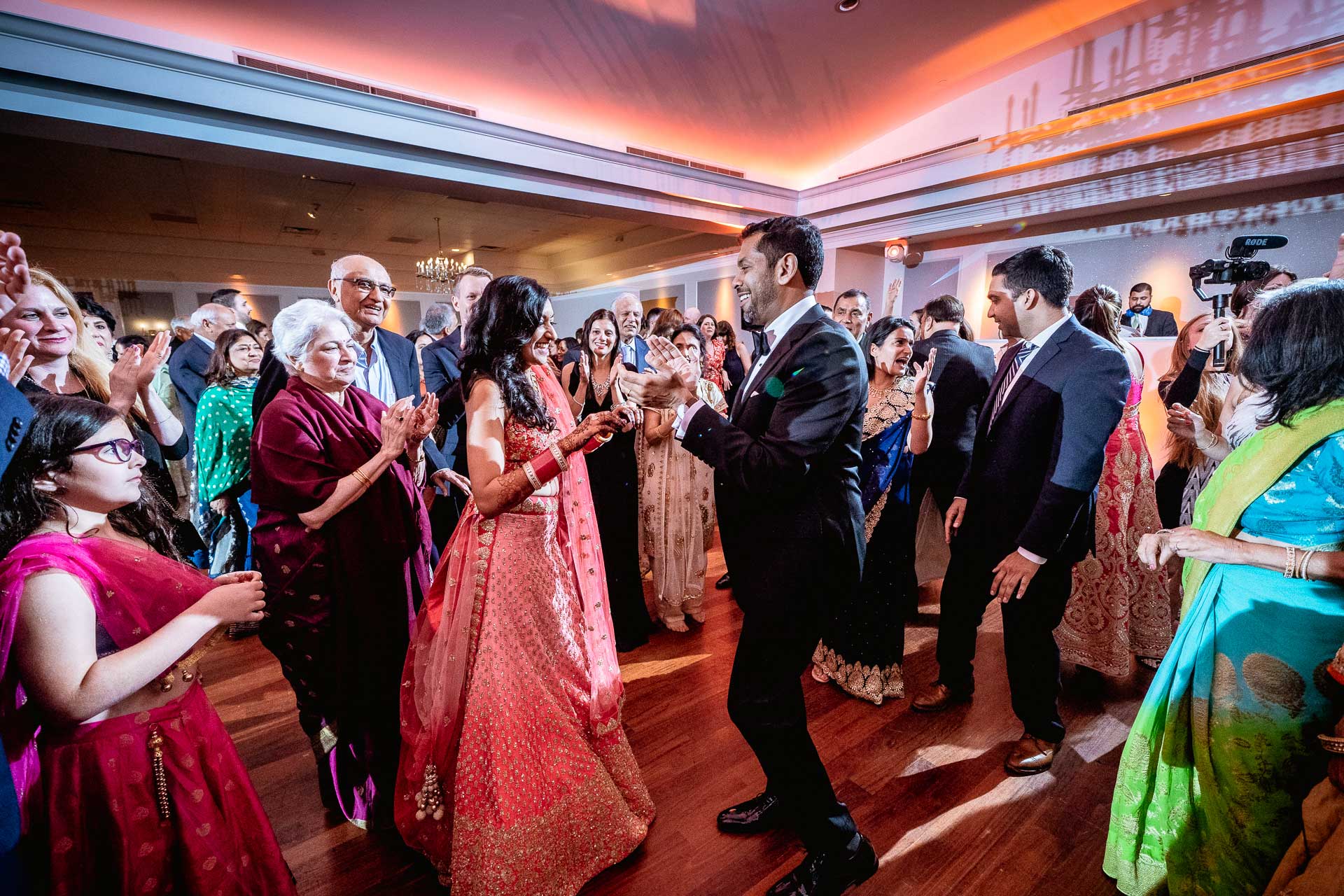 brooklake-Indian-Wedding-photographer-066 A group of people in formal attire dance and celebrate at an indoor NJ Teen Events gathering, with a couple in traditional clothing at the center.