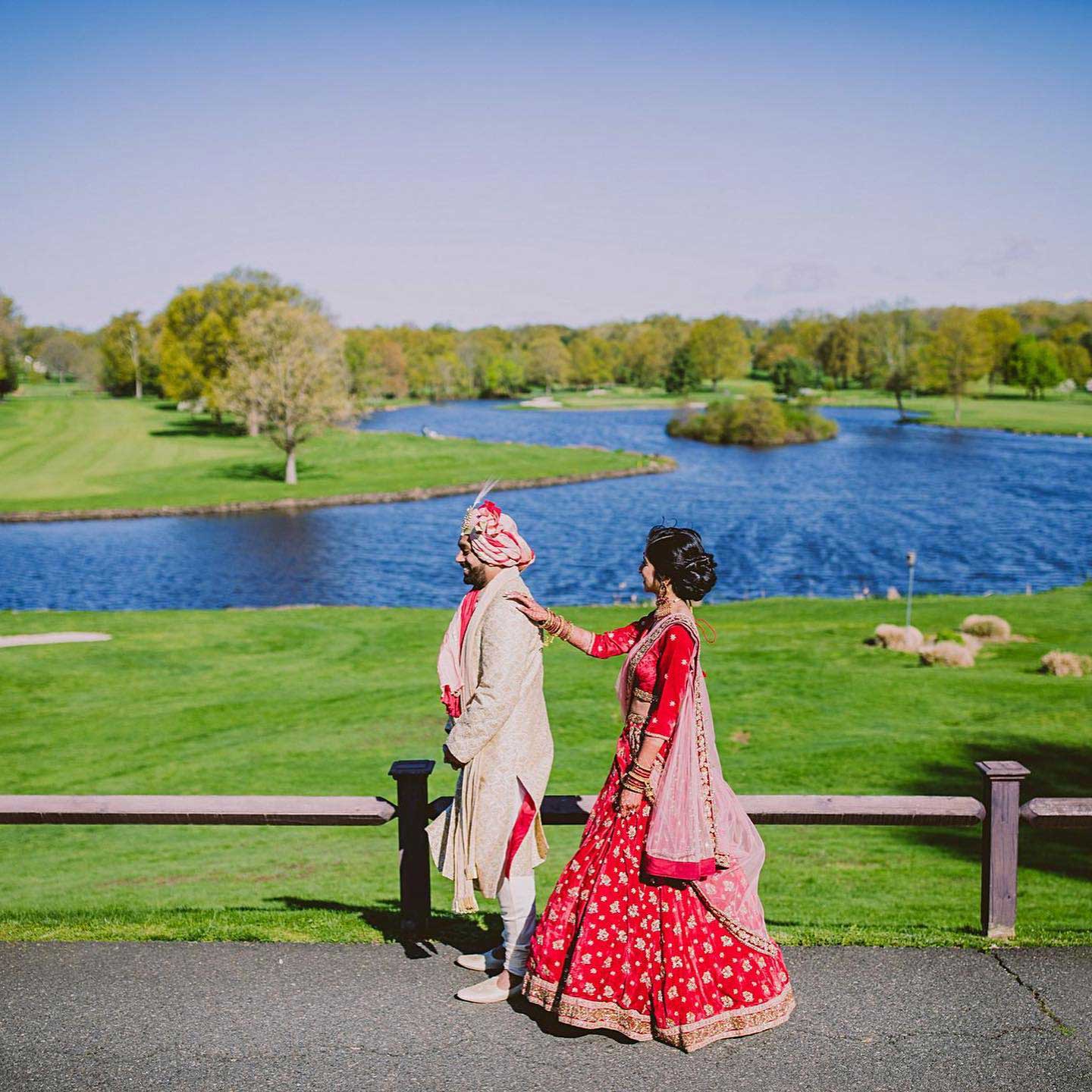 brooklake-bride-groom-first-look-by-lake A bride in a red traditional outfit stands behind a groom in a cream-colored sherwani beside a river on a sunny day, touching his shoulder—capturing the vibrant spirit often seen at NJ Teen Events.