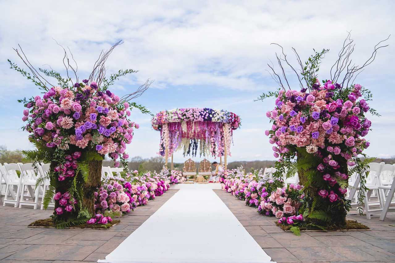 brooklake-colorful-floral-ceremony A wedding aisle with a white runner, lined with rows of chairs and large arrangements of pink and purple flowers, creates an elegant look perfect for special occasions like weddings or NJ Teen Events, leading to a beautifully decorated floral canopy.