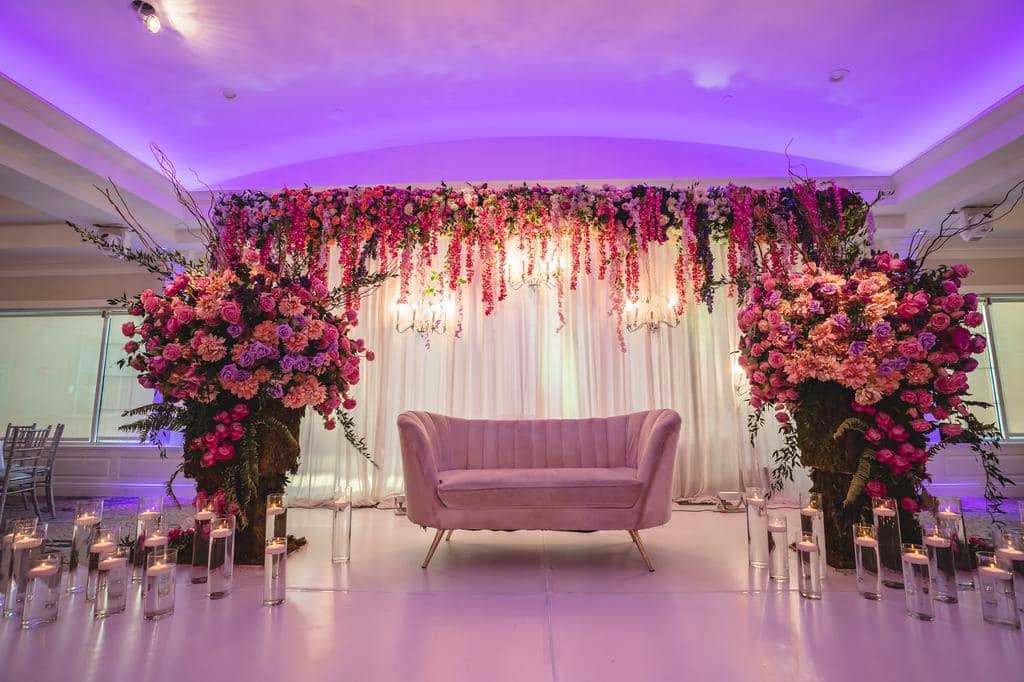 brooklake-colorful-floral-decor-sofa A pink velvet sofa is centered under a floral arch with hanging flowers, perfect for NJ Teen Events, flanked by large flower arrangements and surrounded by lit candles in glass holders.