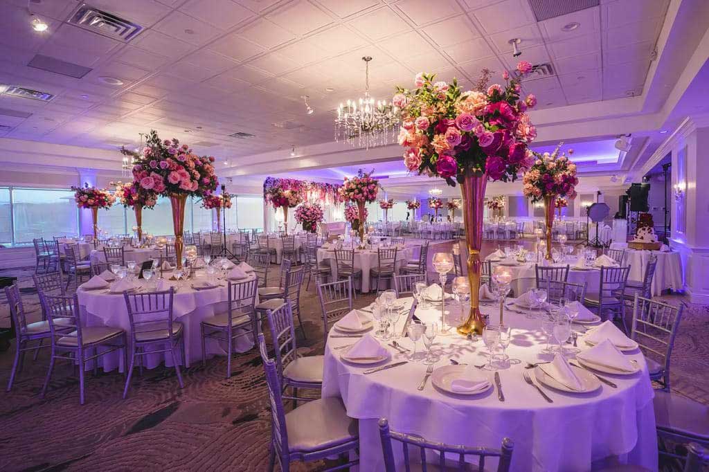 brooklake-colorful-floral-reception A banquet hall set for an event with round tables, white tablecloths, silver chairs, and tall floral centerpieces featuring pink and red flowers under soft purple lighting—perfect for hosting stylish NJ Teen Events.