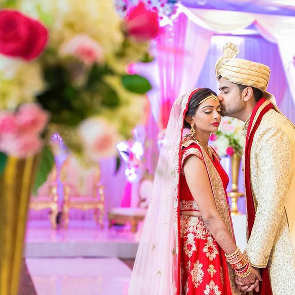 brooklake-indian-bride-groom-colorful-decor A groom kisses the bride’s forehead as they hold hands at a traditional Indian wedding ceremony, with floral decorations in the foreground—capturing a magical moment perfect for NJ Teen Events inspiration.