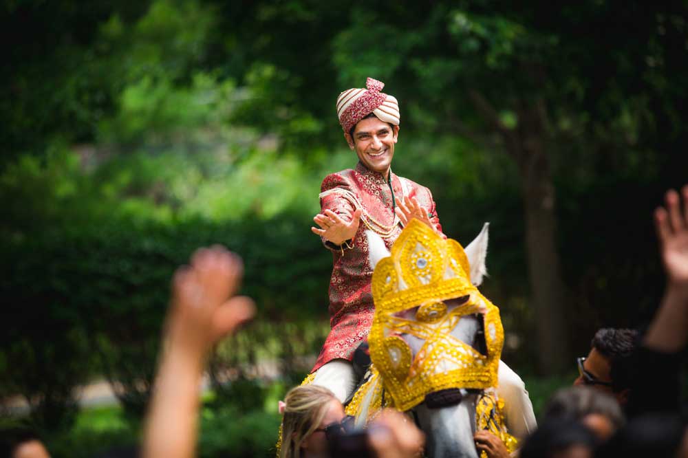brooklake-indian-wedding-arrival-by-horse A man in traditional Indian wedding attire rides a decorated white horse, extending his hand to a crowd with raised hands, outdoors amid greenery—capturing the vibrant energy often seen at NJ Teen Events.