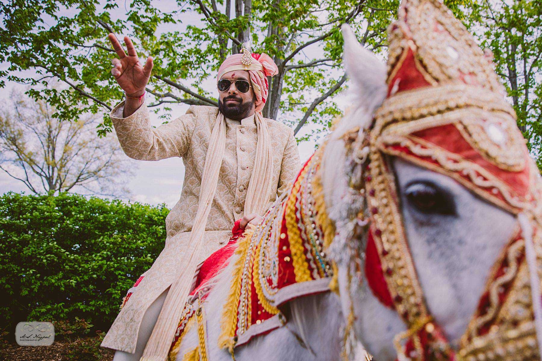brooklake-indian-wedding-arrival-on-horse A man wearing traditional Indian wedding attire and sunglasses rides a decorated white horse, raising his hand in celebration, with green trees in the background—capturing a vibrant moment perfect for NJ Teen Events.