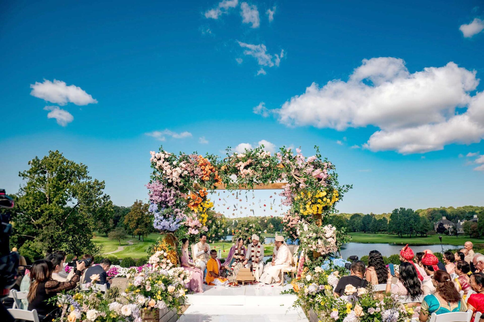 brooklake-indian-wedding-blue-skies-by-lake Outdoor wedding ceremony with guests seated, a floral arch, and a couple under the arch on a sunny day with blue sky and scattered clouds—perfect inspiration for NJ Teen Events seeking elegant outdoor setups.