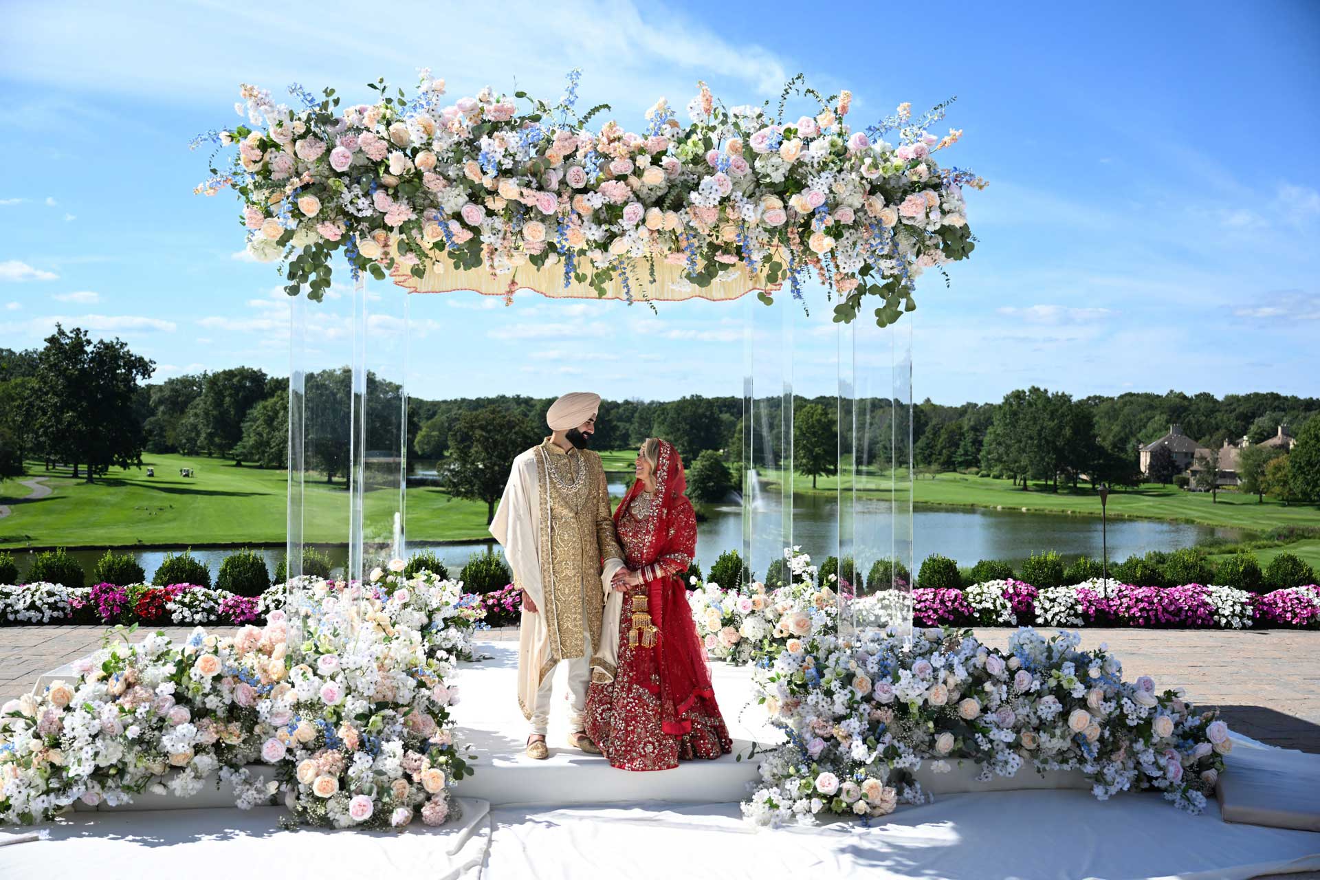 brooklake-indian wedding clear altar A bride and groom in traditional South Asian attire stand under a floral canopy outdoors, surrounded by flower arrangements, as if celebrating one of the elegant NJ Teen Events, with a scenic view of a lake and greenery in the background.