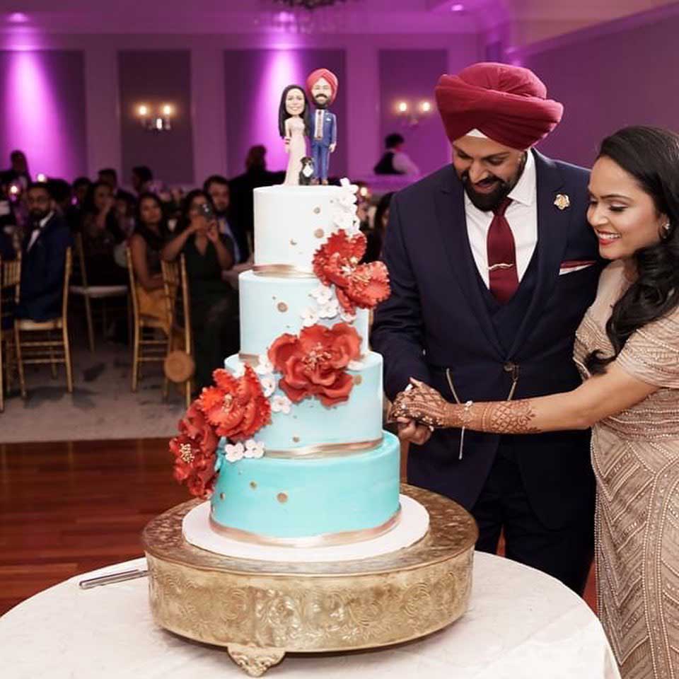 brooklake-indian-wedding-couple-cutting-cake A couple in formal attire cuts a four-tier wedding cake adorned with red flowers and custom figurines at an NJ Teen Events celebration, while guests watch in the background.