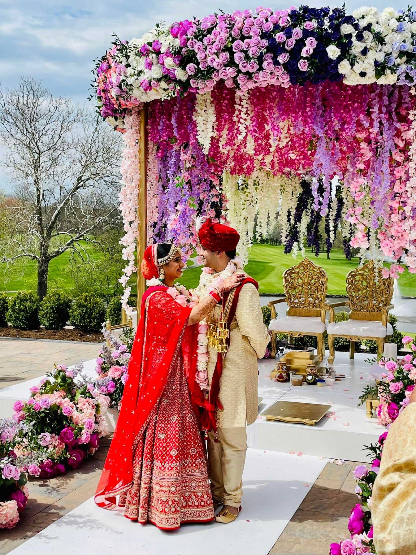 brooklake-indian-wedding-floral-altar A bride and groom in traditional Indian attire stand under a colorful floral wedding canopy outdoors at an NJ Teen Events celebration, surrounded by vibrant floral decorations and ornate chairs.