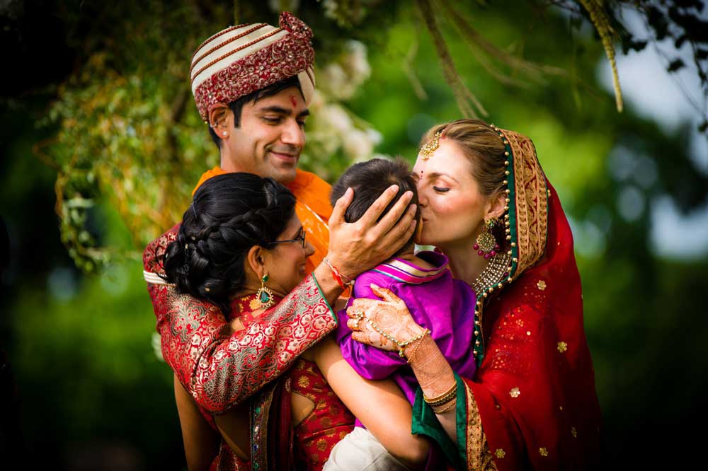 brooklake-indian-wedding-group-kiss-child A couple dressed in traditional South Asian wedding attire embraces and kisses a child amidst lush greenery, capturing a heartwarming moment perfect for NJ Teen Events celebrating culture and family.