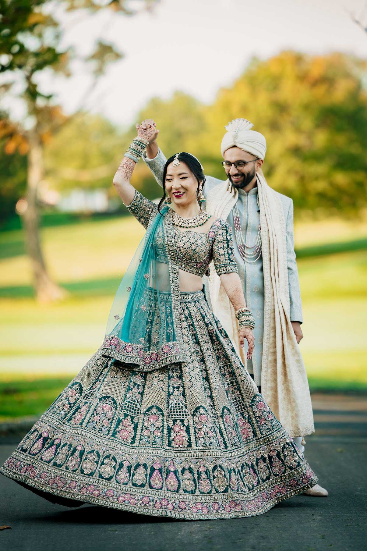 brooklake-south-asian-wedding-bride-groom-dancing A couple in traditional South Asian wedding attire dances outdoors at an NJ Teen Events celebration; the woman wears a detailed embroidered lehenga, while the man dons a turban and sherwani.