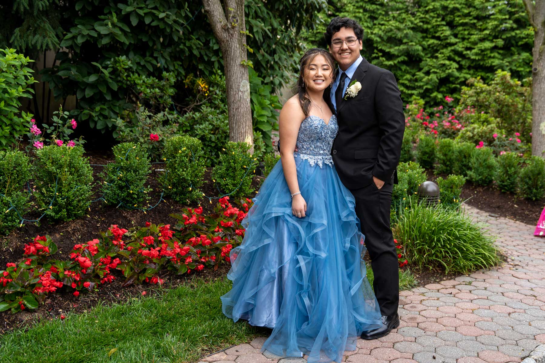 brooklake-teen-couple-at-prom Two people dressed in formal attire, perfect for NJ Bar & Bat Mitzvahs, pose together in a garden with red flowers and green foliage—one in a blue gown, the other in a black suit.