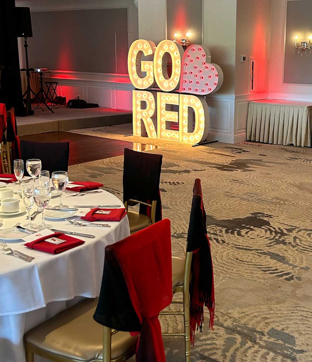 go-red-corporate-event-brooklake A banquet hall set for NJ corporate events features tables ready for guests; a large illuminated "GO RED" sign with a heart brightens the wall, while chairs are dressed in striking red and black covers.