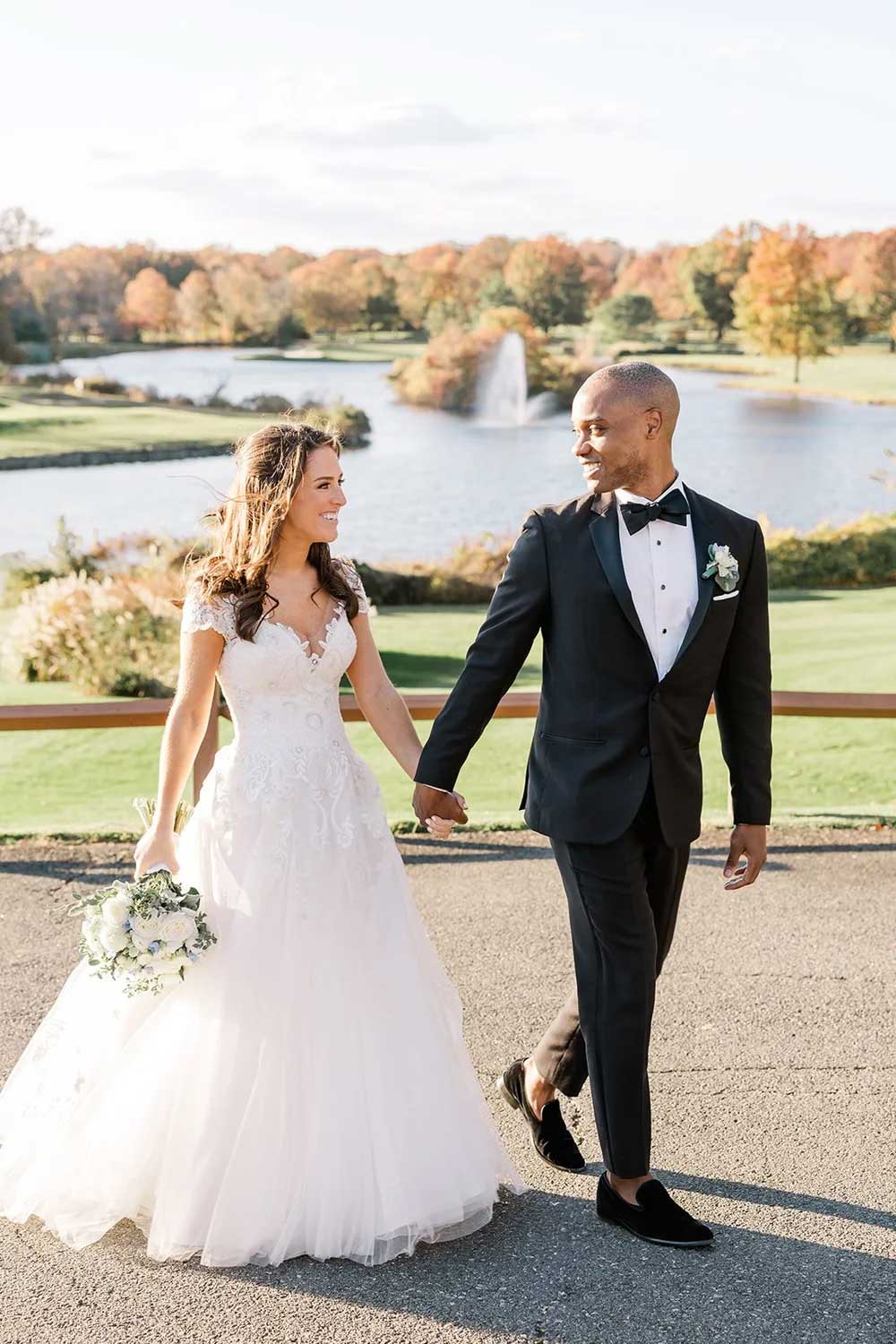 kelly-aaron-walking-lakeside-at-brooklake-wedding A bride and groom walk hand in hand outdoors on a sunny day, dressed in elegant wedding attire, with a lake and trees in the background—perfect for capturing timeless NJ Indian South Asian Weddings moments.