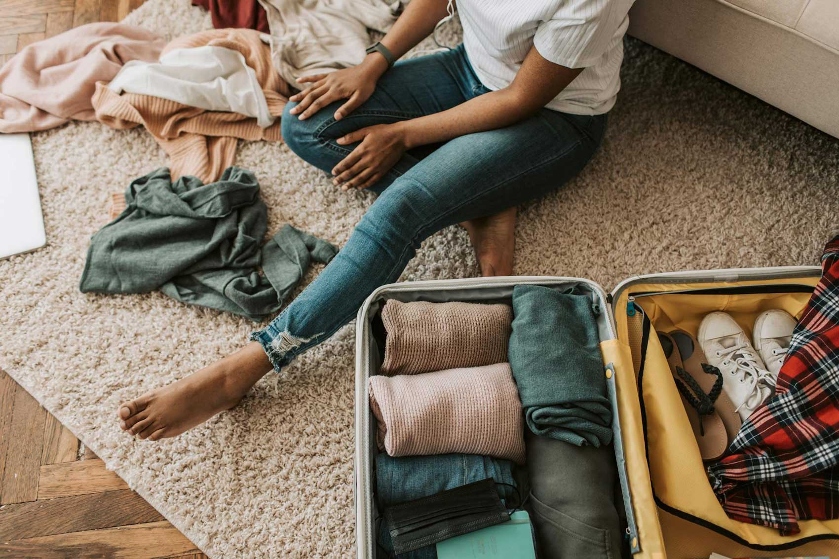packing-for-fall-honeymoon A person sits on a carpeted floor next to an open suitcase, neatly packing clothes and shoes—perhaps preparing for an unforgettable celebration at an NJ wedding & event venue. Scattered clothing items surround them in eager anticipation.