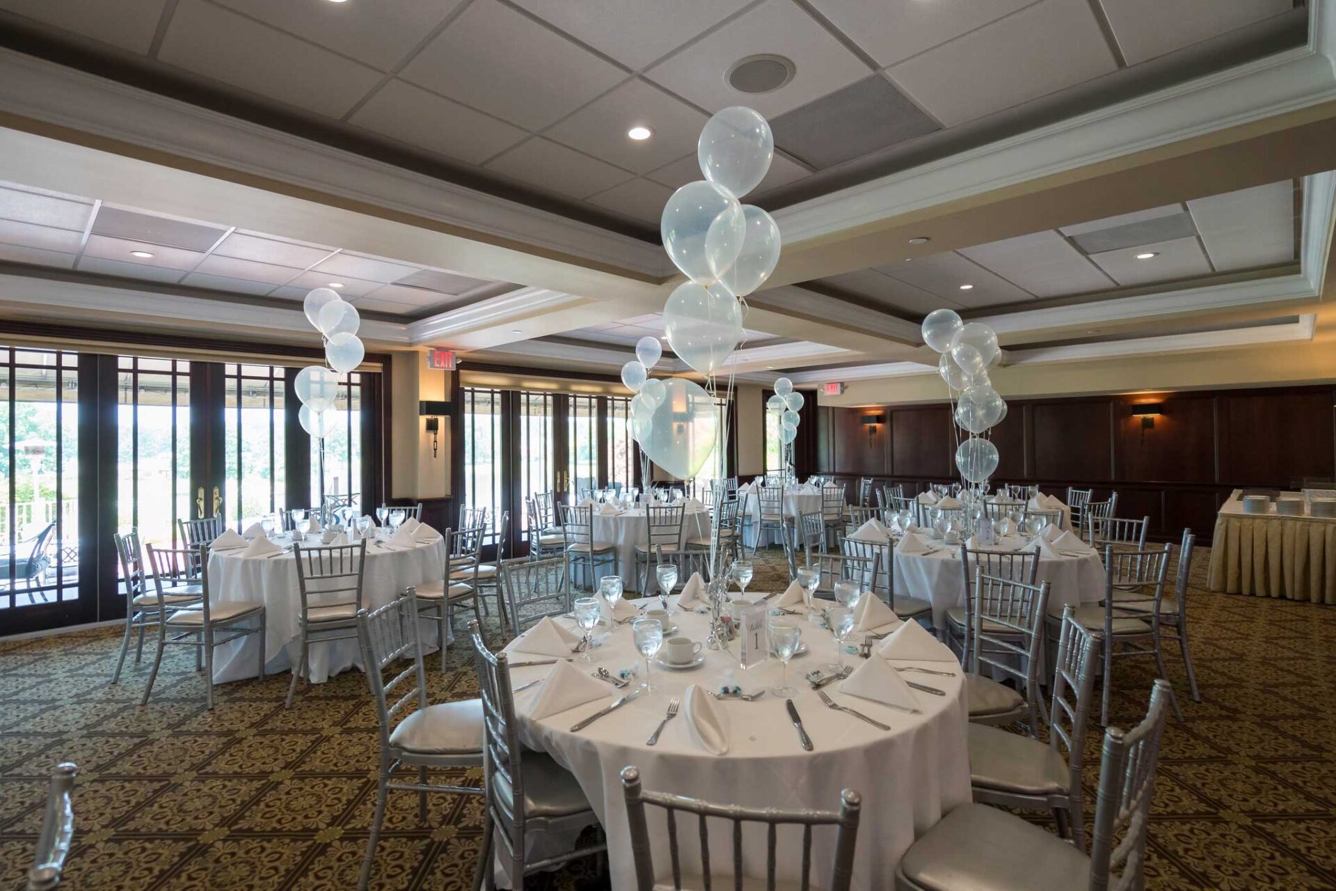 waterford-room-baby-shower-balloons A banquet hall with round tables set for an NJ Teen Events celebration, featuring white tablecloths, silver chairs, and clear balloons as centerpieces.