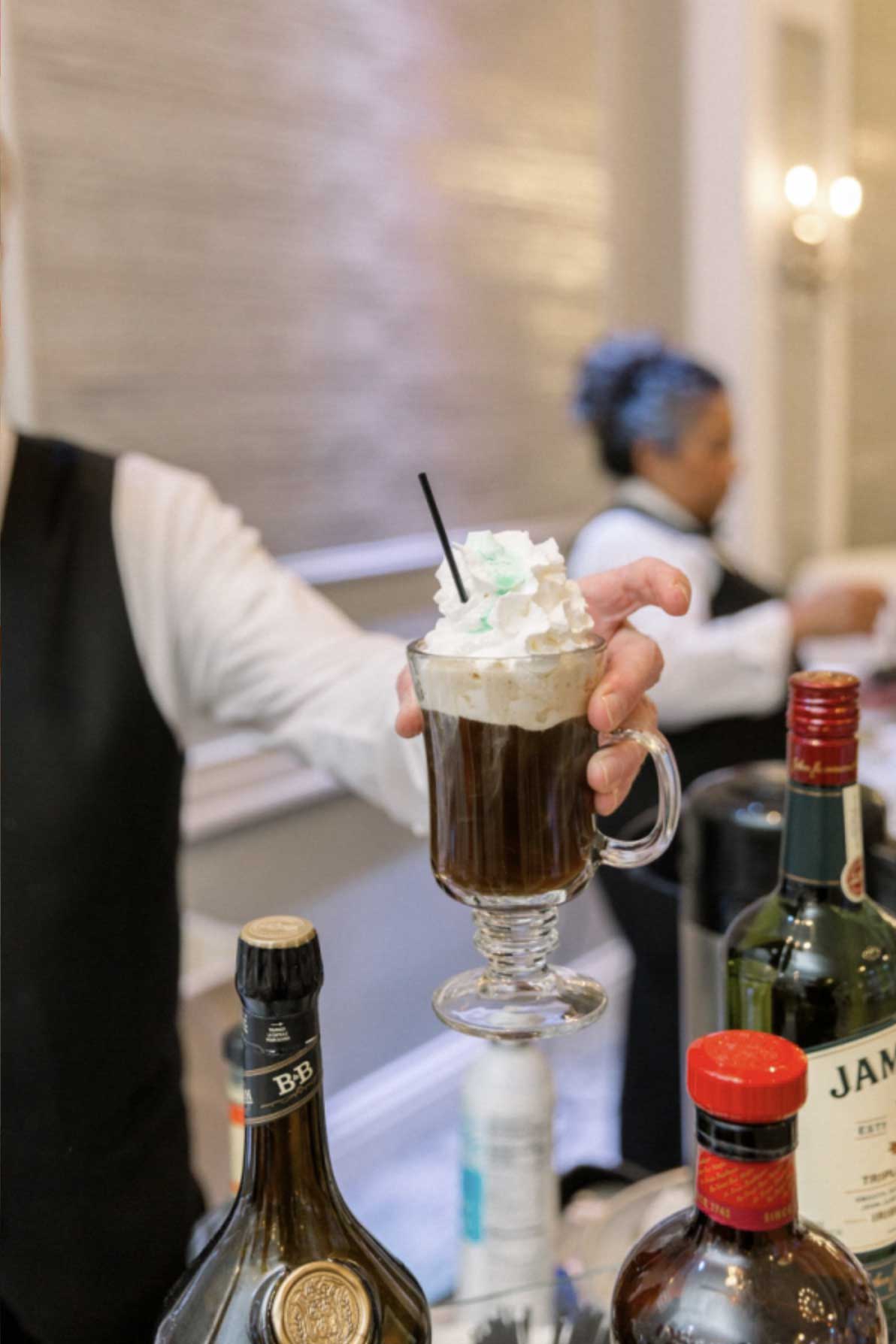 brooklake-bar-beverage-drinks A server holds a glass mug of Irish coffee topped with whipped cream, with various bottles of liquor on the counter—perfect for warming up guests at NJ corporate events.