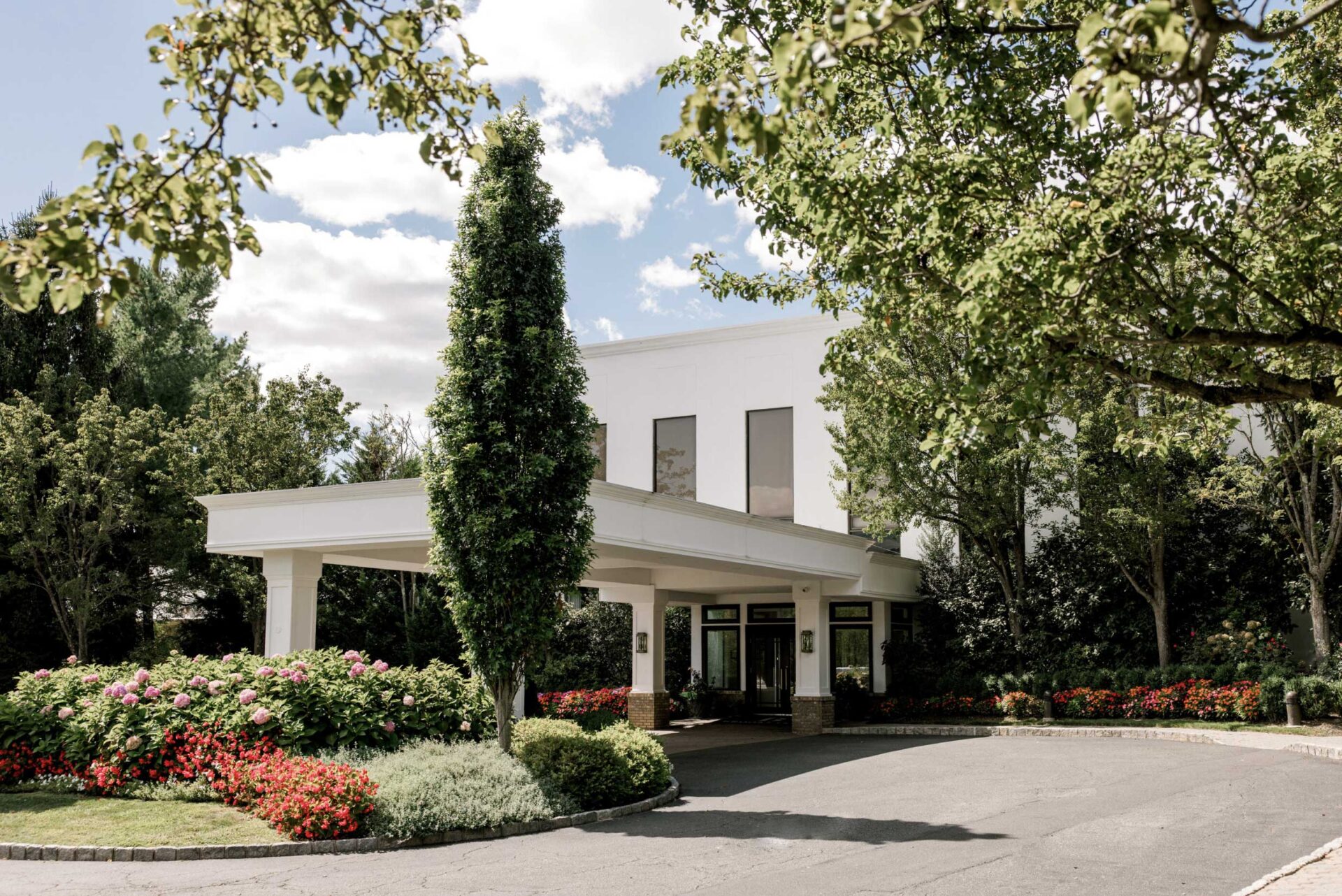 brooklake-entry A white building entrance with large windows, perfect for NJ Indian South Asian Weddings, is surrounded by manicured gardens, flower beds, and trees under a partly cloudy sky.