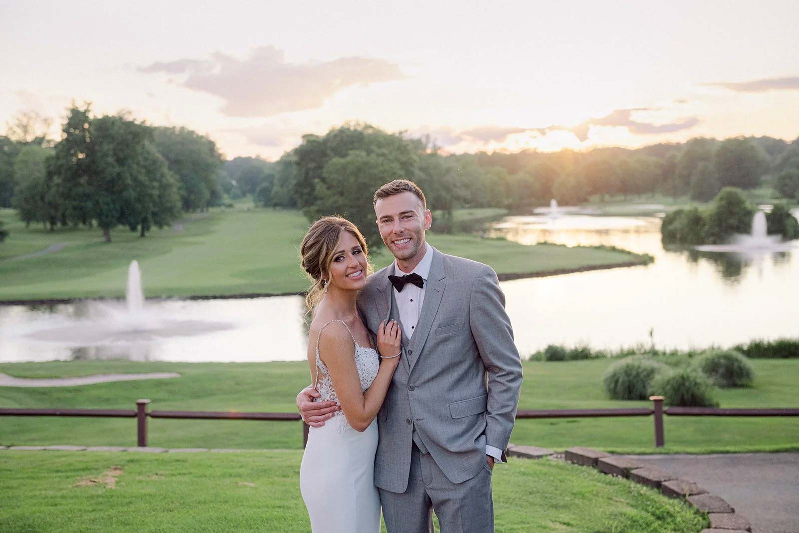 bride-and-groom-on-lawn-outside-at-brooklake-wedding-ceremony A couple poses in formal wedding attire outdoors at sunset, standing on grass with a pond, fountains, trees, and a golf course in the background—an ideal setting for unforgettable Wedding & Event Cuisine.