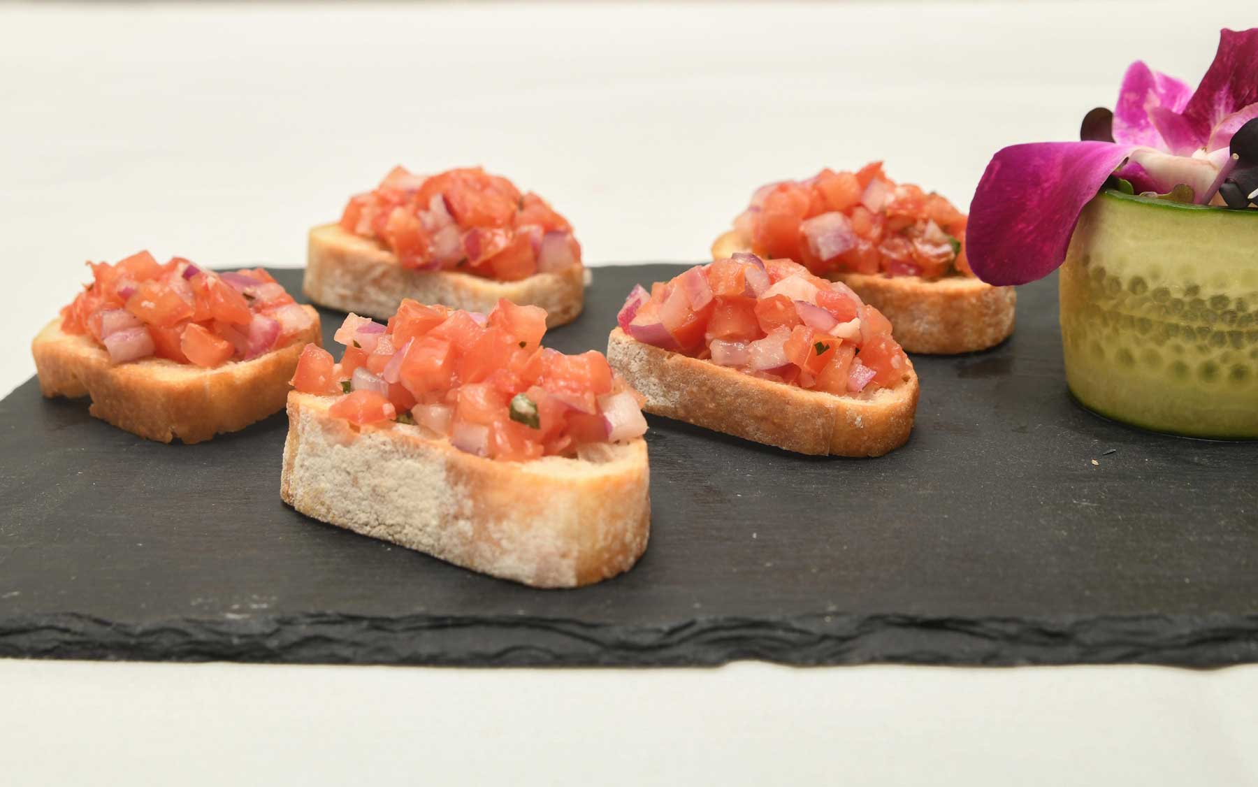 appetizer-bruschetta Five pieces of bruschetta with diced tomatoes and onions on a slate platter, next to a cucumber slice garnished with a purple flower—a perfect touch for Wedding & Event Cuisine.