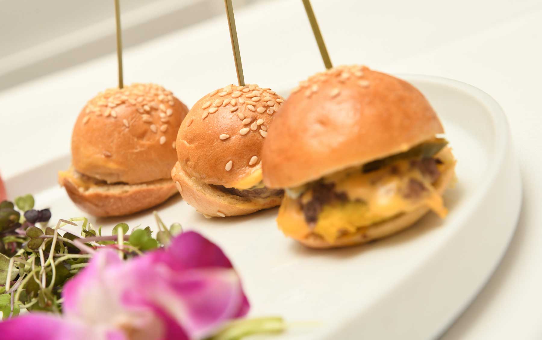 brooklake-appetizer-sliders Three mini cheeseburgers with sesame seed buns are served on a white plate, garnished with microgreens and a purple flower—perfect for sophisticated Wedding & Event Cuisine.