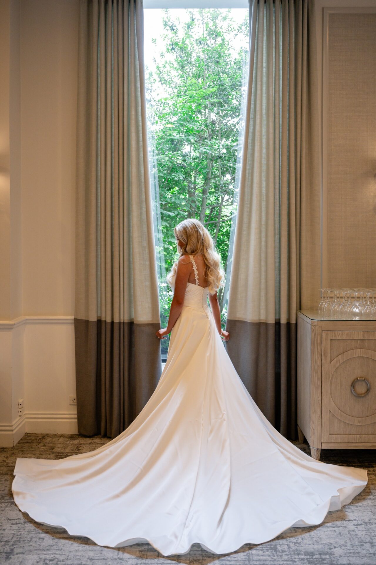 brooklake-bride-at-window-634052656_1519173820214683_1005042521967752648_n A woman in a white wedding dress stands facing a large window with curtains, holding the sides, as greenery from an NJ wedding venue is visible outside.