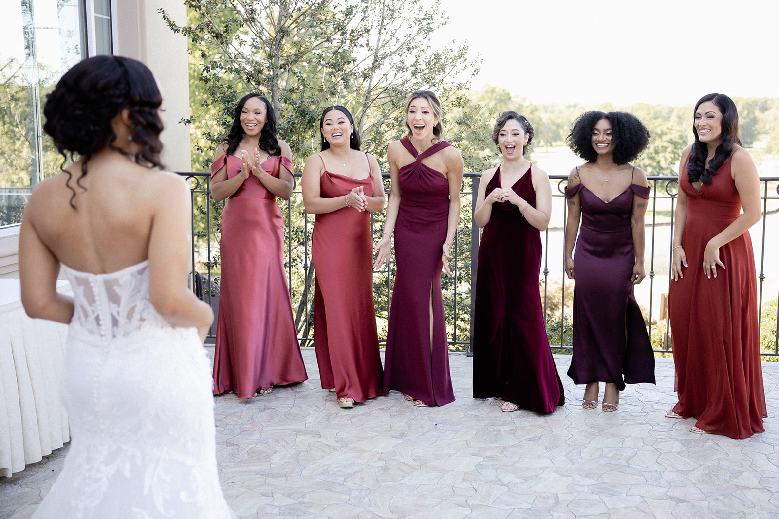 brooklake-bride-with-bridesmaids-balcony-624551845_1507038254761573_3158605581400221009_n A bride in a white wedding dress faces six bridesmaids in burgundy dresses, who are smiling and reacting with excitement on an outdoor patio at a stunning NJ wedding venue.