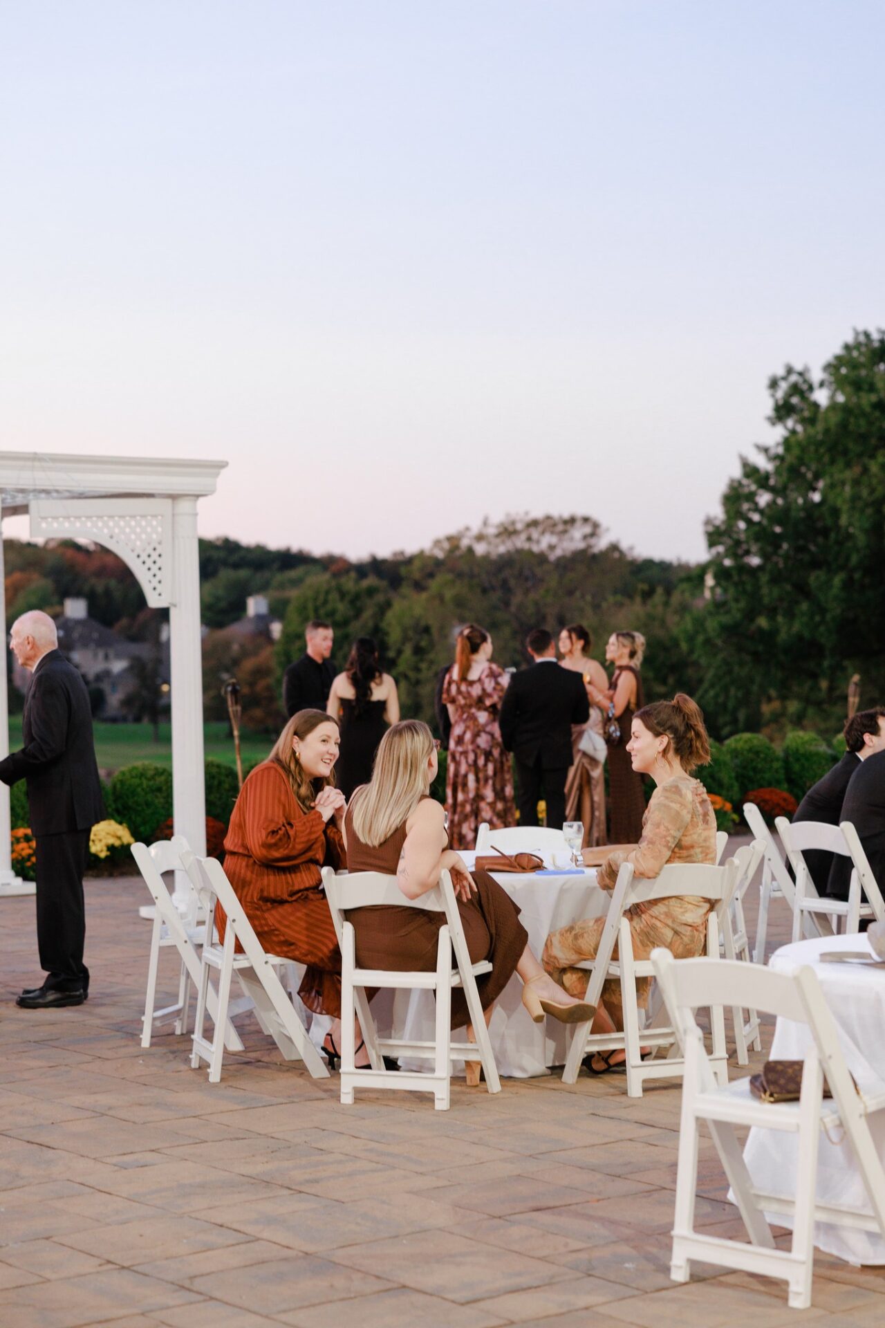 brooklake-guests-647342361_1538496411615757_2027417102535829745_n Guests sit and talk at a table with white chairs outdoors while others mingle in the background during an event at sunset at an elegant NJ wedding venue.