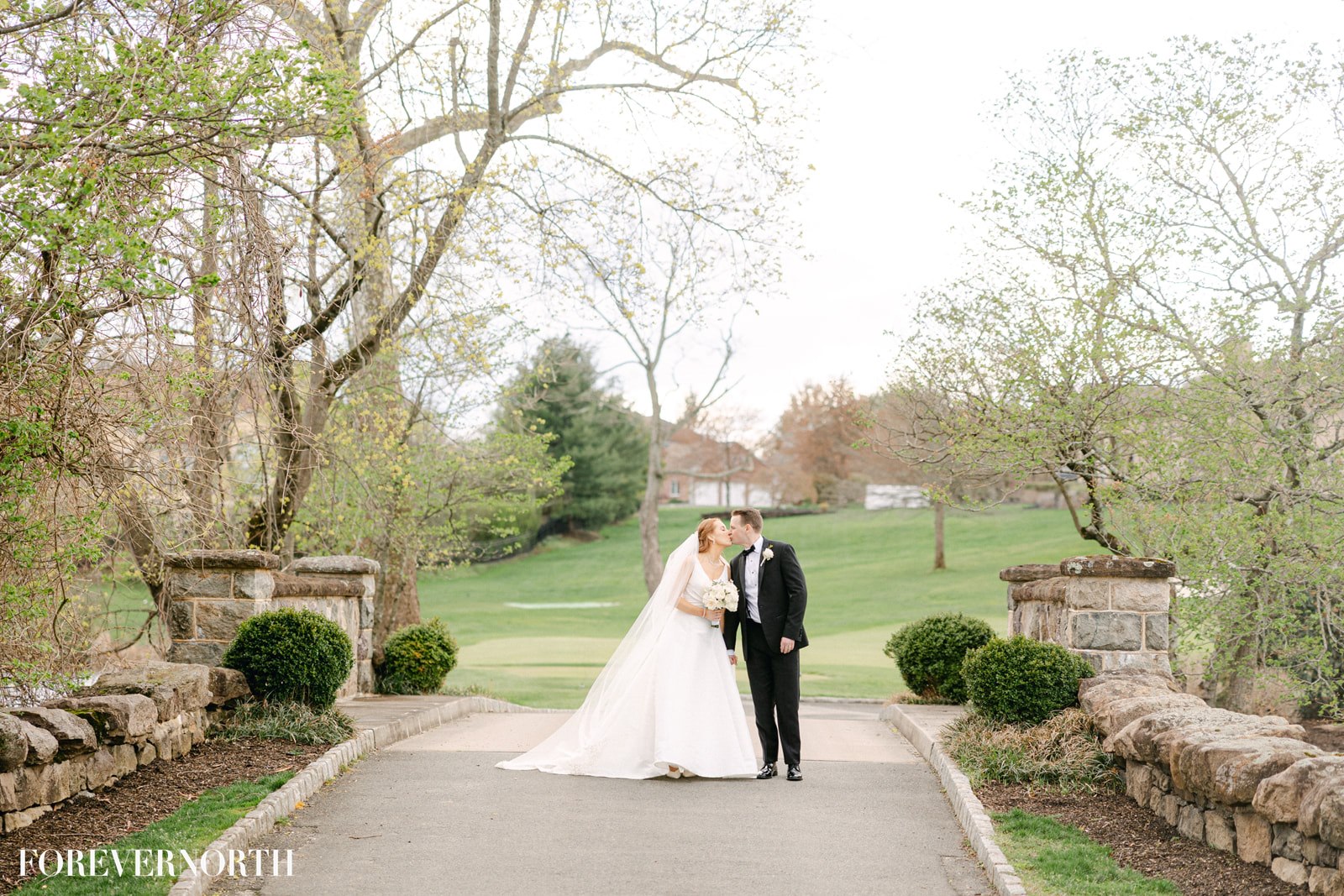 brooklake-property-bride-groom-651177759_1549919767140088_434674223511651376_n A bride and groom stand on a stone bridge at an elegant NJ wedding venue, kissing, surrounded by trees and greenery on a golf course. The bride wears a white dress; the groom wears a black tuxedo.